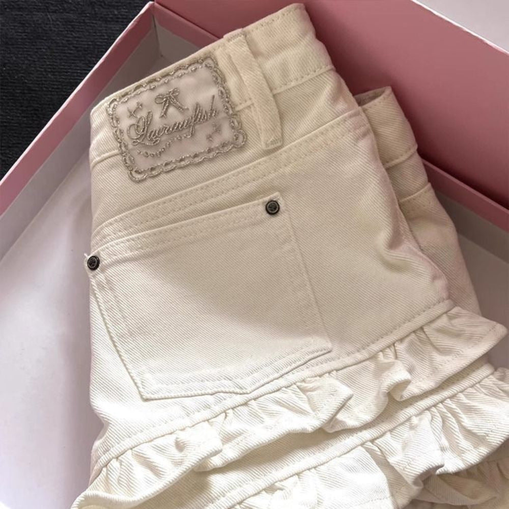 Close-up back view of cream-colored denim shorts featuring a multi-tiered ruffled hem, back pocket, and elegant embroidered 'Sugarwish' brand label, presented in a pink gift box. Cute and feminine fashion detail for Boutique Jolie.