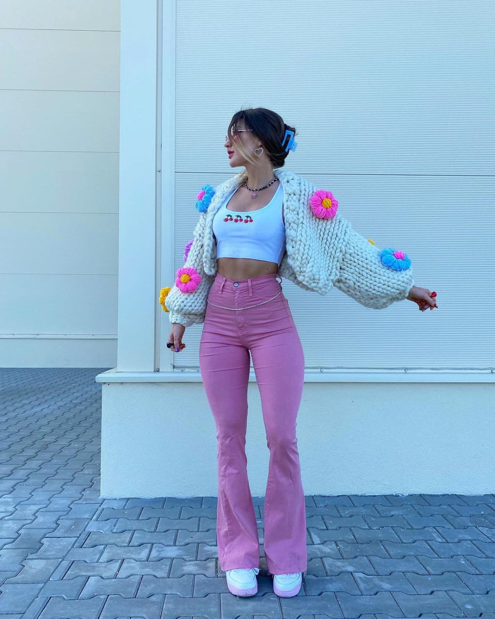 Model wearing a fun oversized white cardigan decorated with multicolored pom-poms over a white crop top, paired with pink pants and white sneakers. Confident full-body pose with arms slightly outstretched in an outdoor setting against a modern white architectural wall.