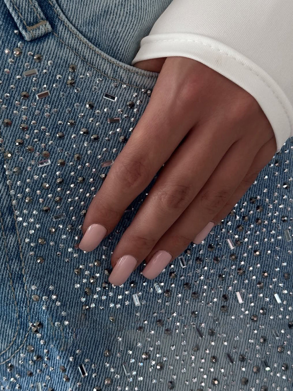 Close-up of hand with light pink manicured nails on rhinestone embellished blue denim fabric.