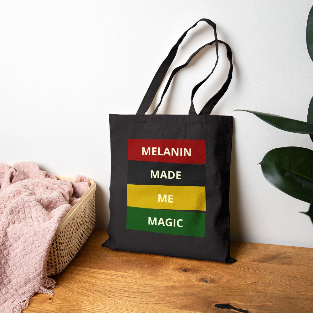 Black canvas tote bag with "MELANIN MADE ME MAGIC" graphic in red, black, yellow, and green blocks on wooden table