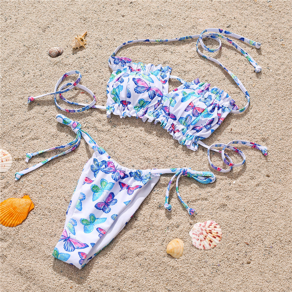 White colorful butterfly print bikini on sand – trendy ruffled swimwear set with ties, beach product flat lay.