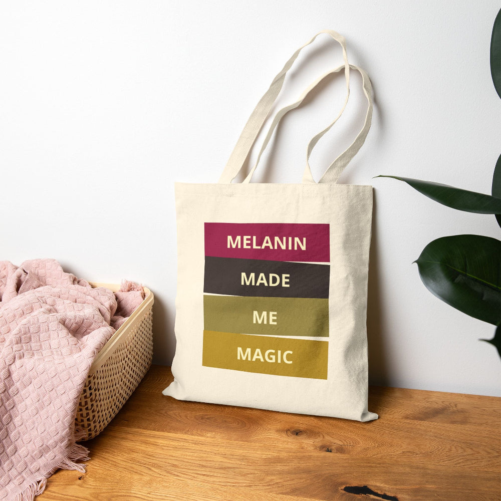 Natural canvas tote bag with "MELANIN MADE ME MAGIC" colorful block text displayed on wooden table with pink throw and plant.