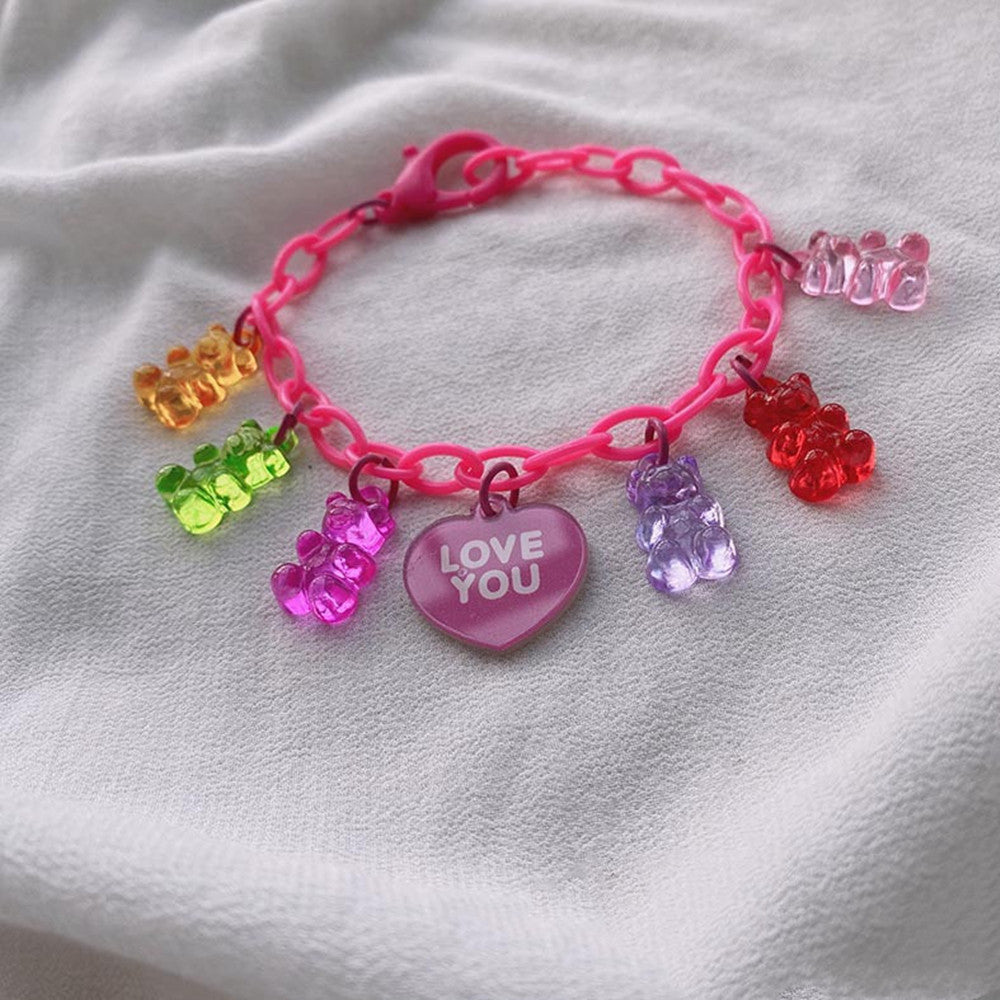 Pink gummy bear charm bracelet featuring colorful translucent gummy bear charms in rainbow colors and a pink heart-shaped pendant engraved with "LOVE YOU", displayed on white fabric.