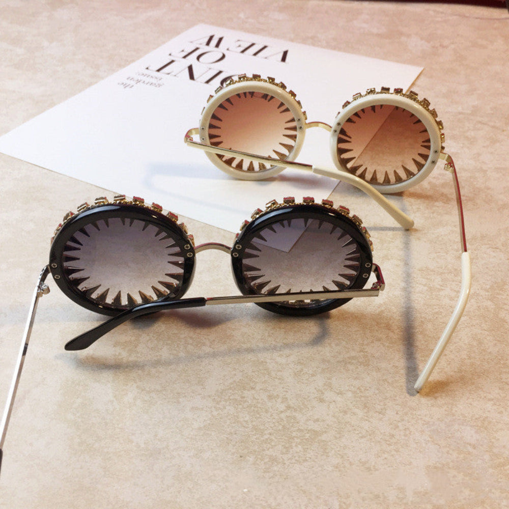 Black and white round steampunk sunglasses with spiked rims and gradient lenses, styled on a beige surface.