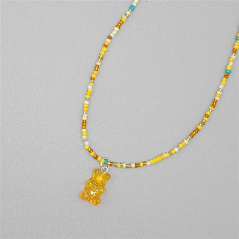 Sunny yellow beaded necklace with a cheerful mix of yellow, white, golden brown, and turquoise seed beads paired with a translucent yellow gummy bear pendant. Playful single-strand design laid flat on a light gray background, highlighting its bright and fun summer vibe.