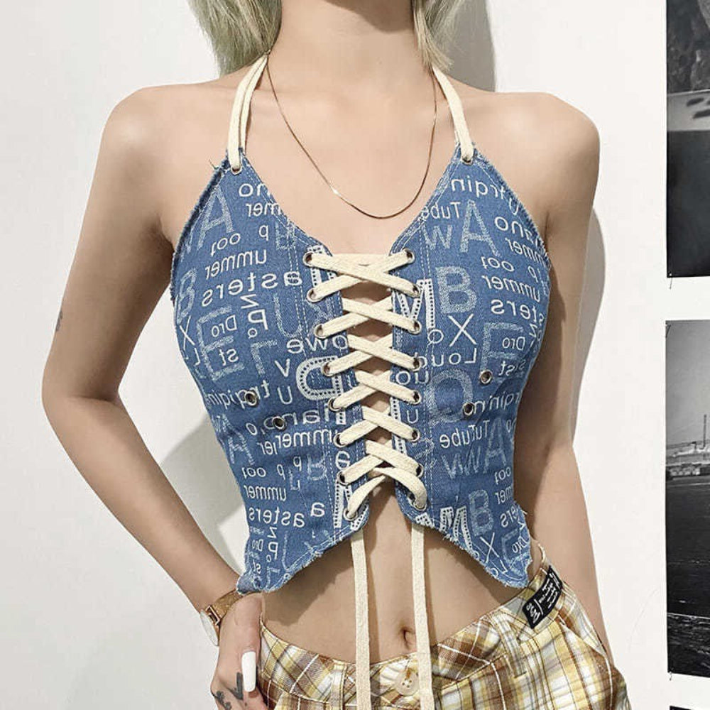 Front view of a woman wearing a blue number and letter print halter crop top with white lace-up corset front, adjustable halter ties, and long dangling strings. Features a deep V-neckline and raw hem details, paired with plaid pants.