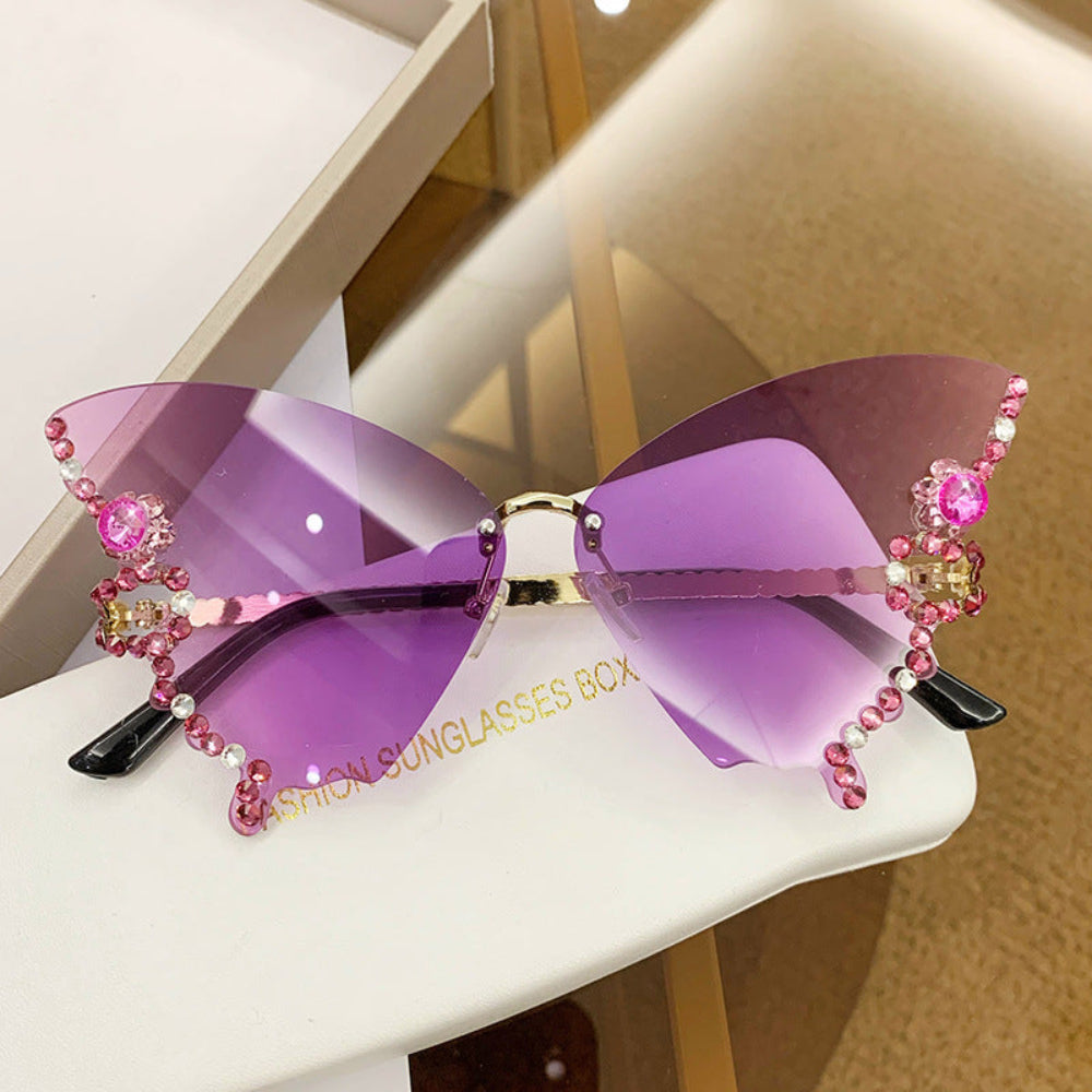 Rimless butterfly sunglasses with purple-pink lenses and pink crystal wing details, displayed on a white glasses case.