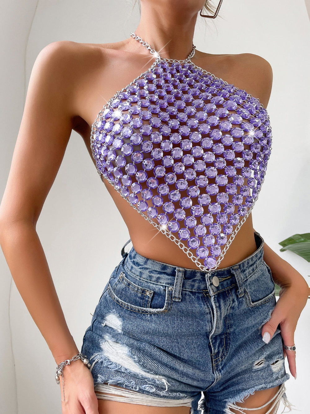 Purple rhinestone crystal halter crop top with diamond-shaped embellishment and chain straps, paired with ripped blue denim shorts, worn by a model.