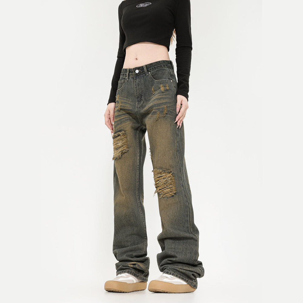 Front view of a model wearing dark greyish-green wide-leg baggy jeans with heavy distressing, large ripped holes, and frayed edges on the thighs and knees. The jeans have a relaxed oversized fit that stacks at the ankles, paired with a black long-sleeve crop top and chunky white sneakers with gum soles.