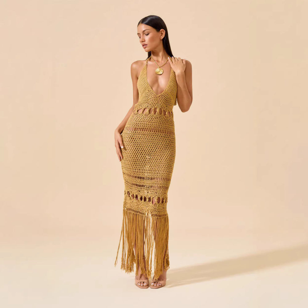 Woman modeling full-length mustard yellow crochet halter dress with fringe hem and waist cutouts against beige studio background.