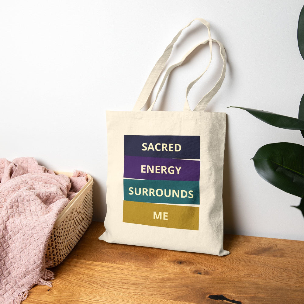 Canvas tote bag with "SACRED ENERGY SURROUNDS ME" spiritual quote in colorful stacked blocks (navy, purple, teal, gold), styled on wooden surface with pink blanket and plant.