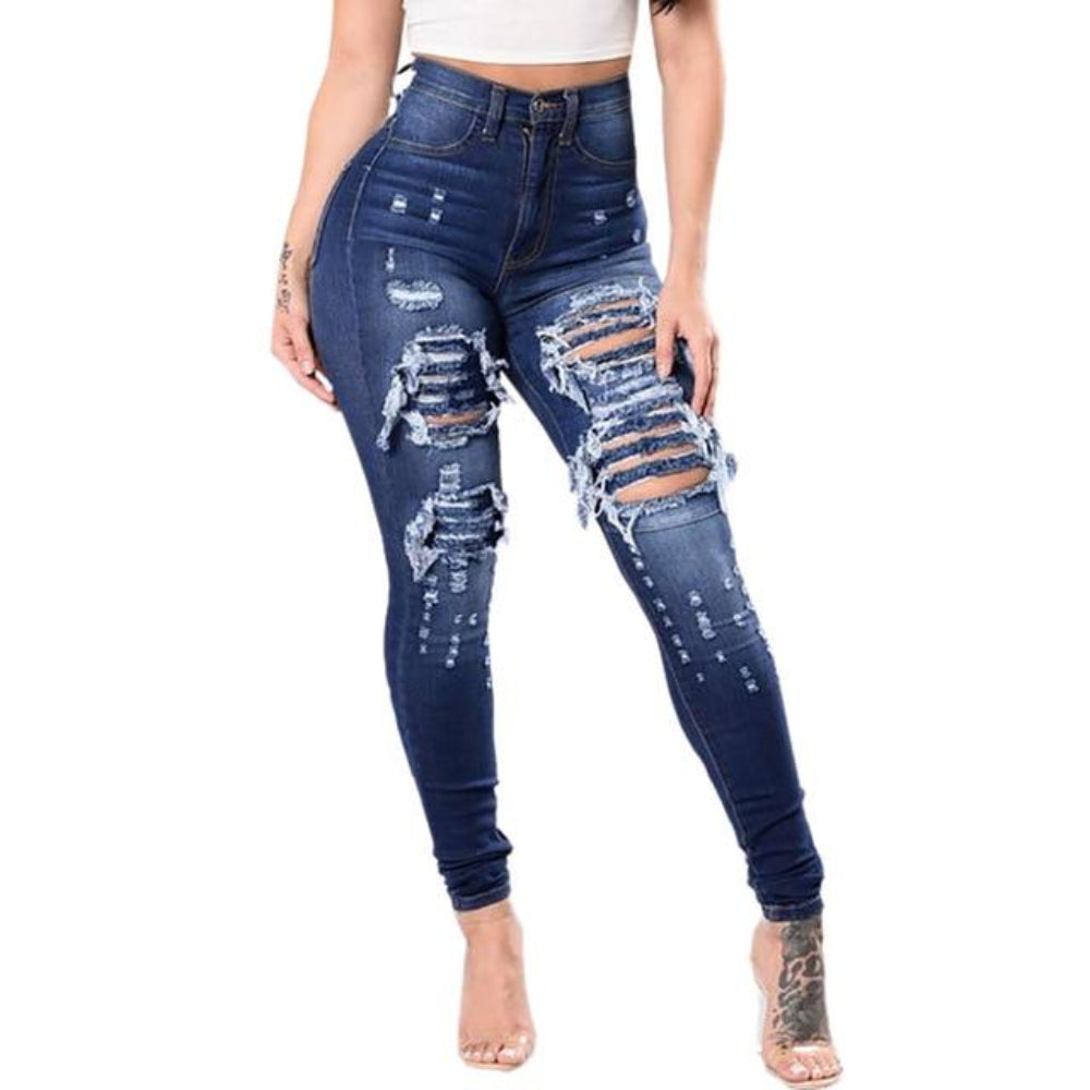 Front view of a woman wearing high-waisted dark blue skinny jeans with heavy distressing and large ripped holes with frayed edges on the thighs and knees. The jeans feature a figure-hugging, stretch fit that accentuates curves, paired with a white crop top and clear high-heeled sandals. Visible tattoos on her arms and lower leg.