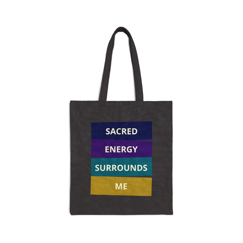 Black canvas tote bag with "SACRED ENERGY SURROUNDS ME" spiritual quote in colorful stacked blocks (navy, purple, teal, gold) on dark fabric against white background.