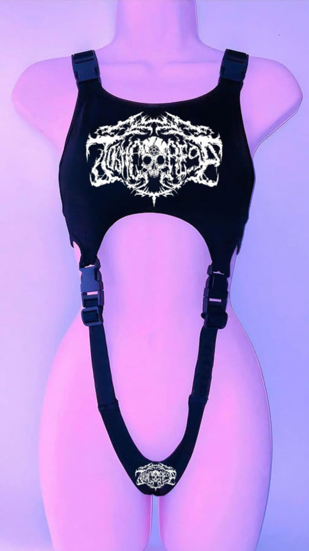 Black harness-style crop top on a mannequin with adjustable buckle straps and side cut-outs. Features a bold white graphic print with skull and stylized text on the chest, plus a smaller matching graphic at the bottom, shown under purple lighting.