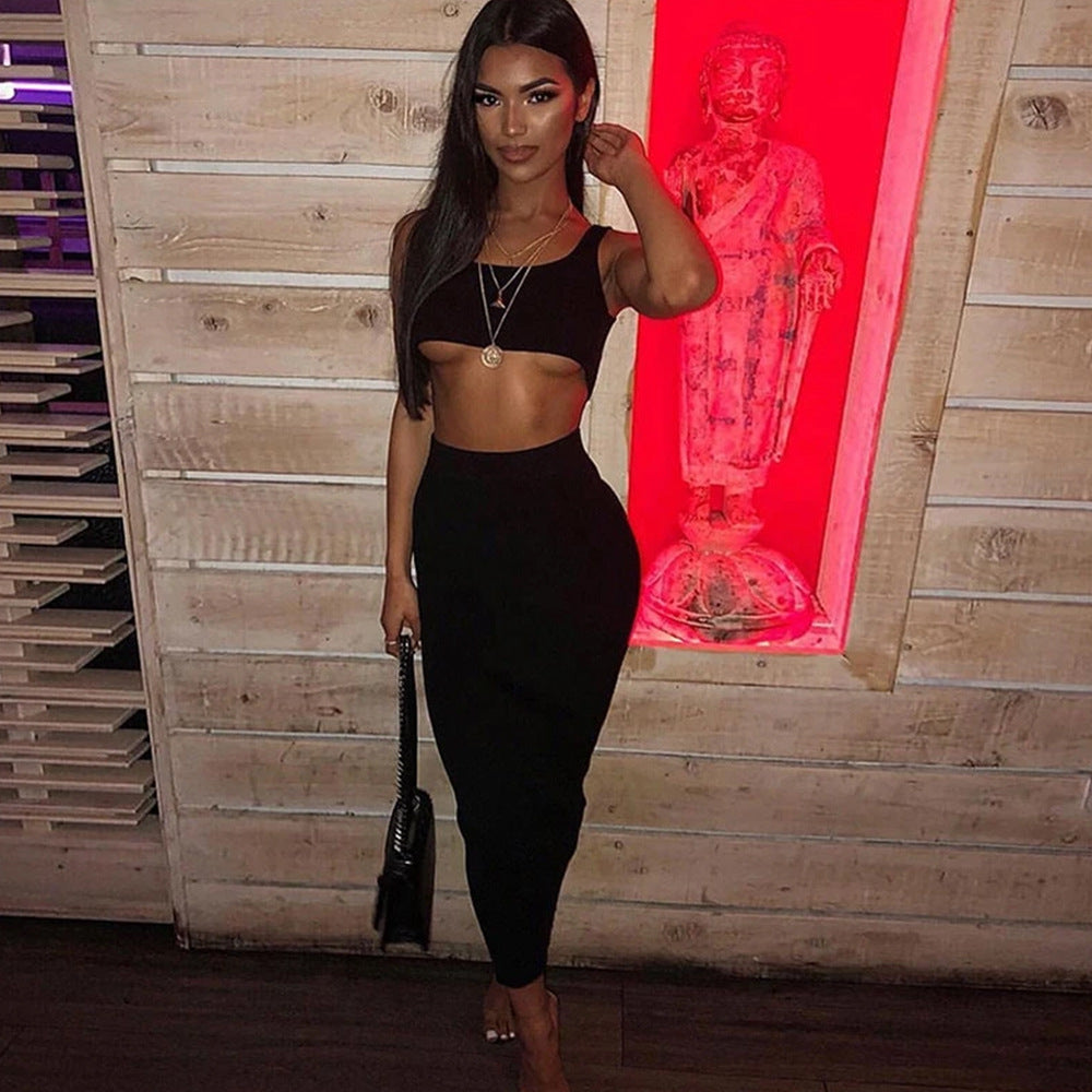 Model in a form-fitting black two-piece outfit (sleeveless crop top and high-waisted midi skirt) standing in a stylish pose with one hand in her hair, holding a black handbag. Shot in front of a glowing red neon sign on a rustic wooden wall background.