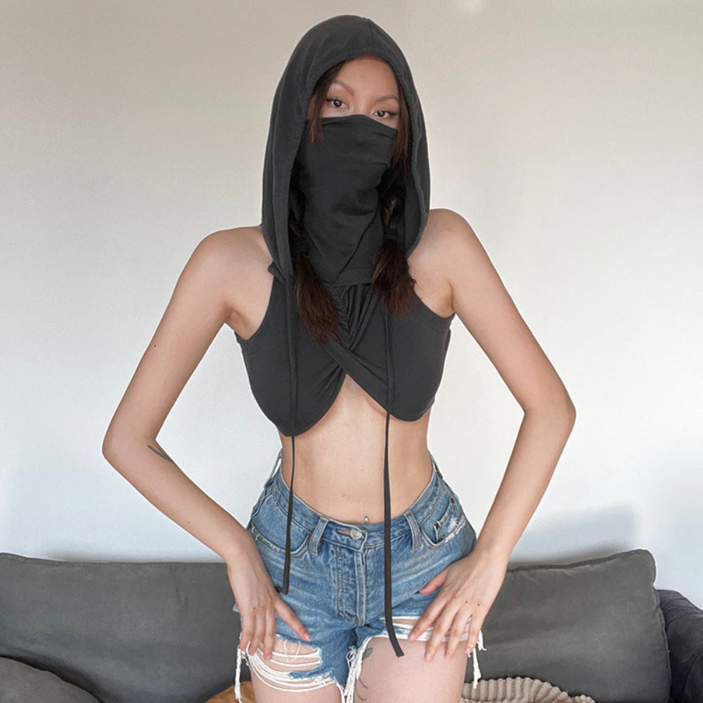 Woman wearing a black hooded crop top with a balaclava-style hood covering most of her face, twisted and draped fabric at the chest, and long adjustable ties. Styled with light-wash distressed denim shorts, posing with hands on hips.