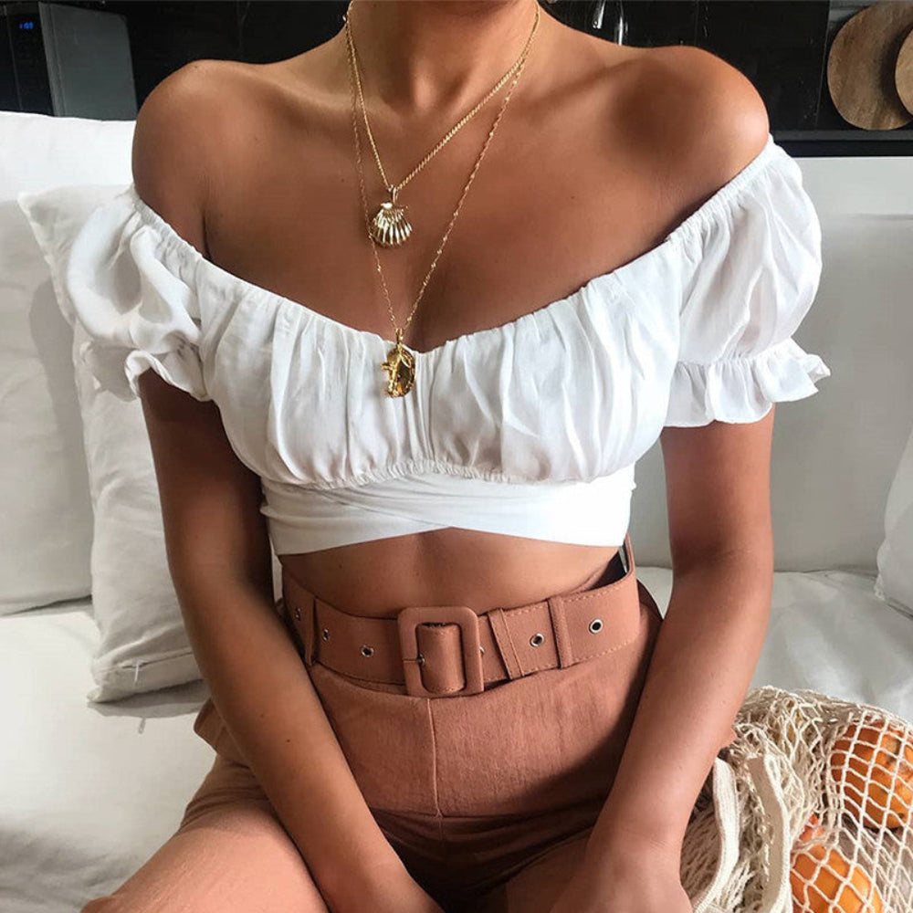 White off-shoulder ruched crop top with puff sleeves, worn by a model with layered gold necklaces and brown pants.