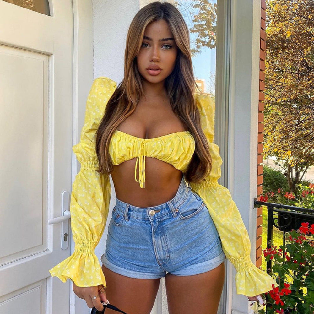 Woman with long wavy brown hair modeling a bright yellow floral print off-shoulder crop top with voluminous puff sleeves, ruched elastic bodice, and front tie detail. Paired with high-waisted light-wash denim shorts, posing confidently at a sunlit doorway with greenery in the background.