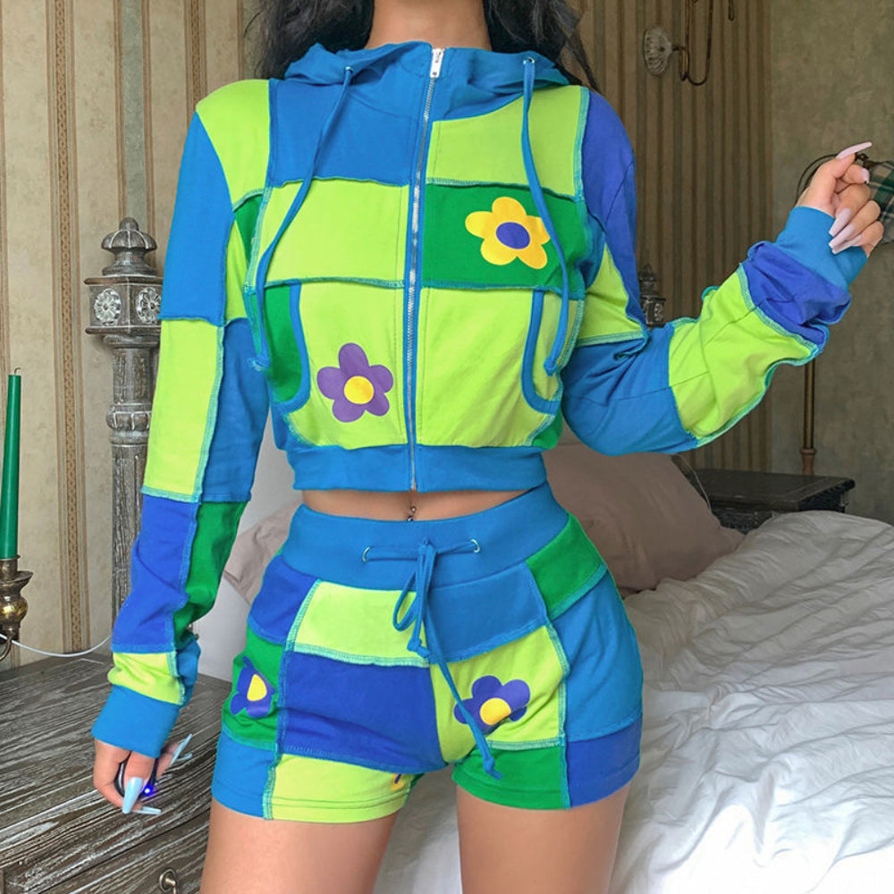 Woman modeling a vibrant patchwork cropped zip-up hoodie and matching shorts set in bright blue, lime green, and green color-block panels. The hoodie features a hood with drawstrings, front zipper, patch pockets, and colorful daisy flower appliqués, while the shorts have a drawstring waist and coordinating flower details. Styled with a visible belly button piercing.