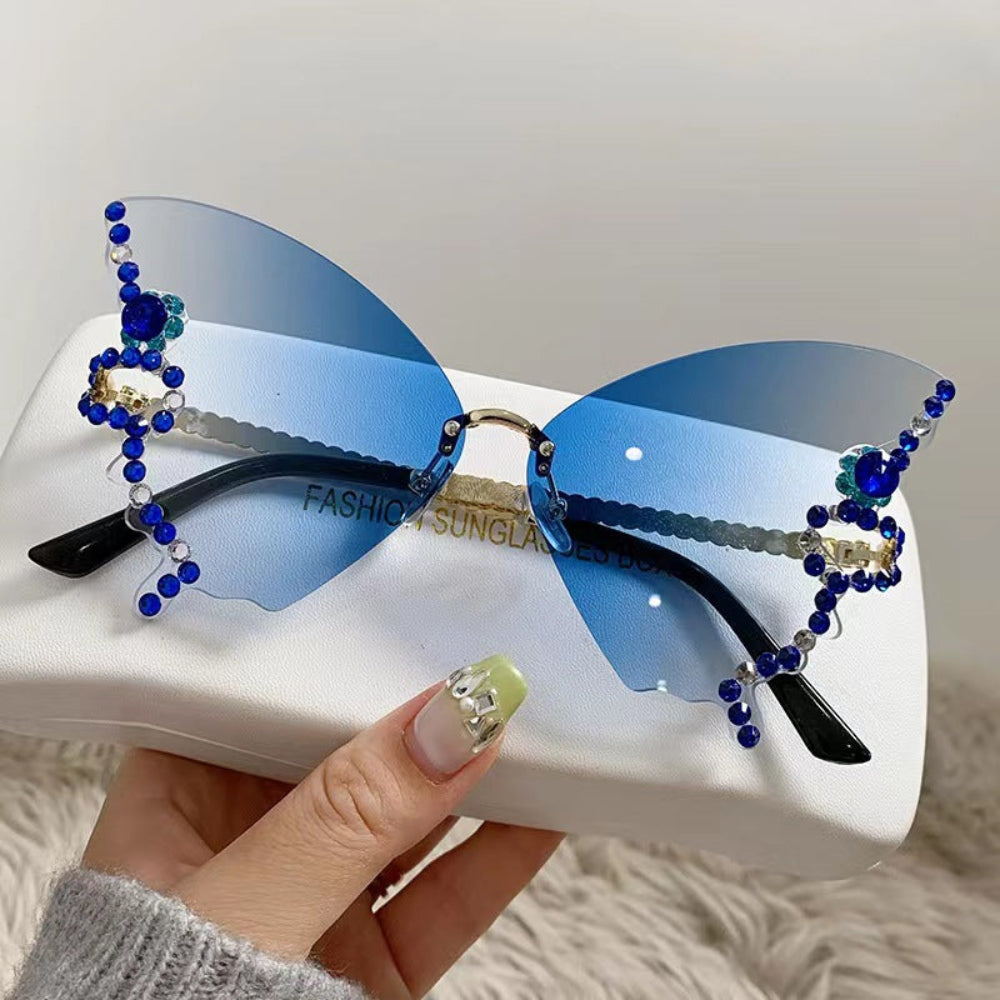 Rimless butterfly sunglasses with blue gradient lenses and blue crystal wing details, held on a white glasses case.