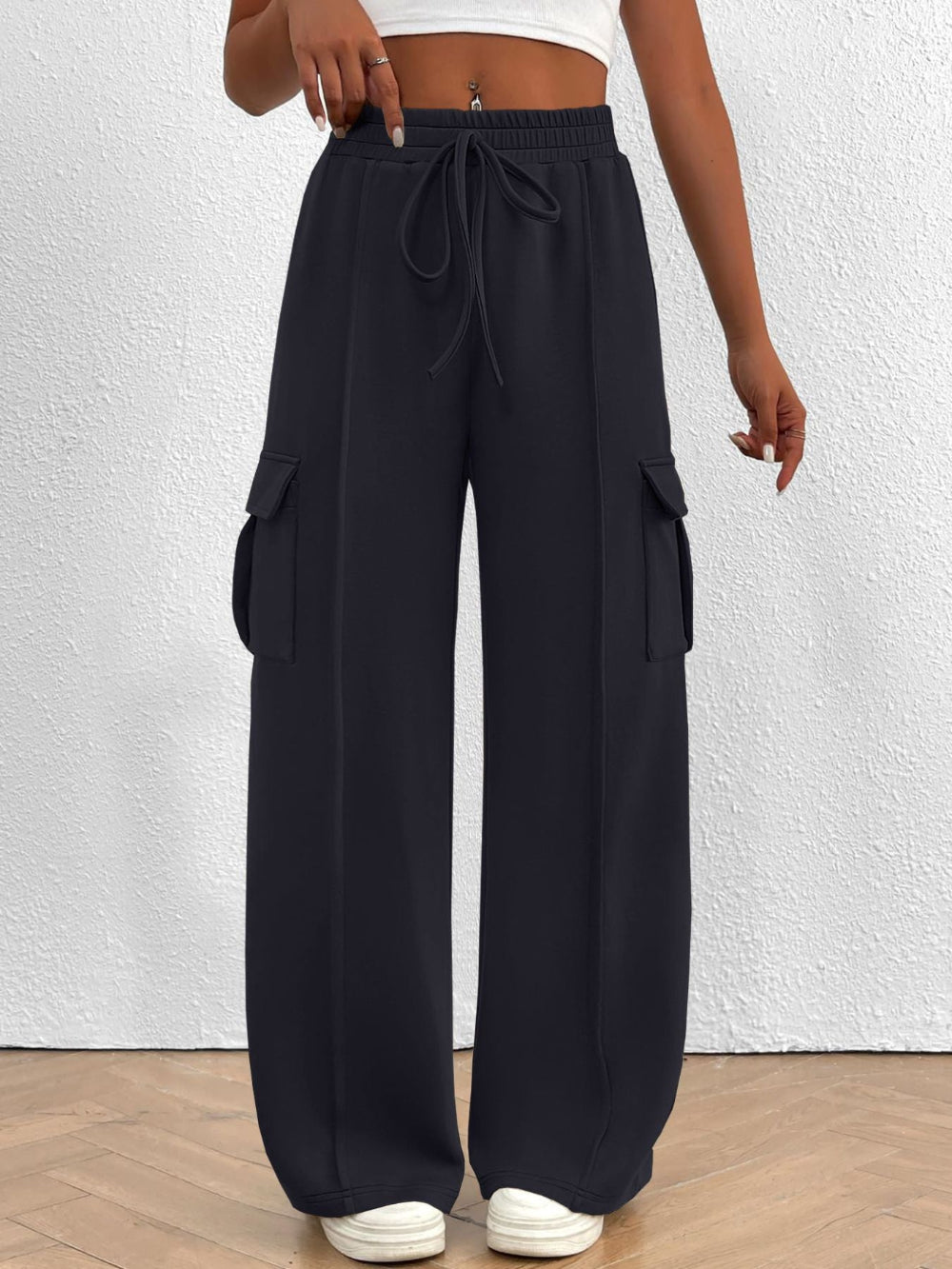 Woman wearing black wide leg cargo pants with drawstring waist and large side pockets paired with a white top and white sneakers against a textured white wall background