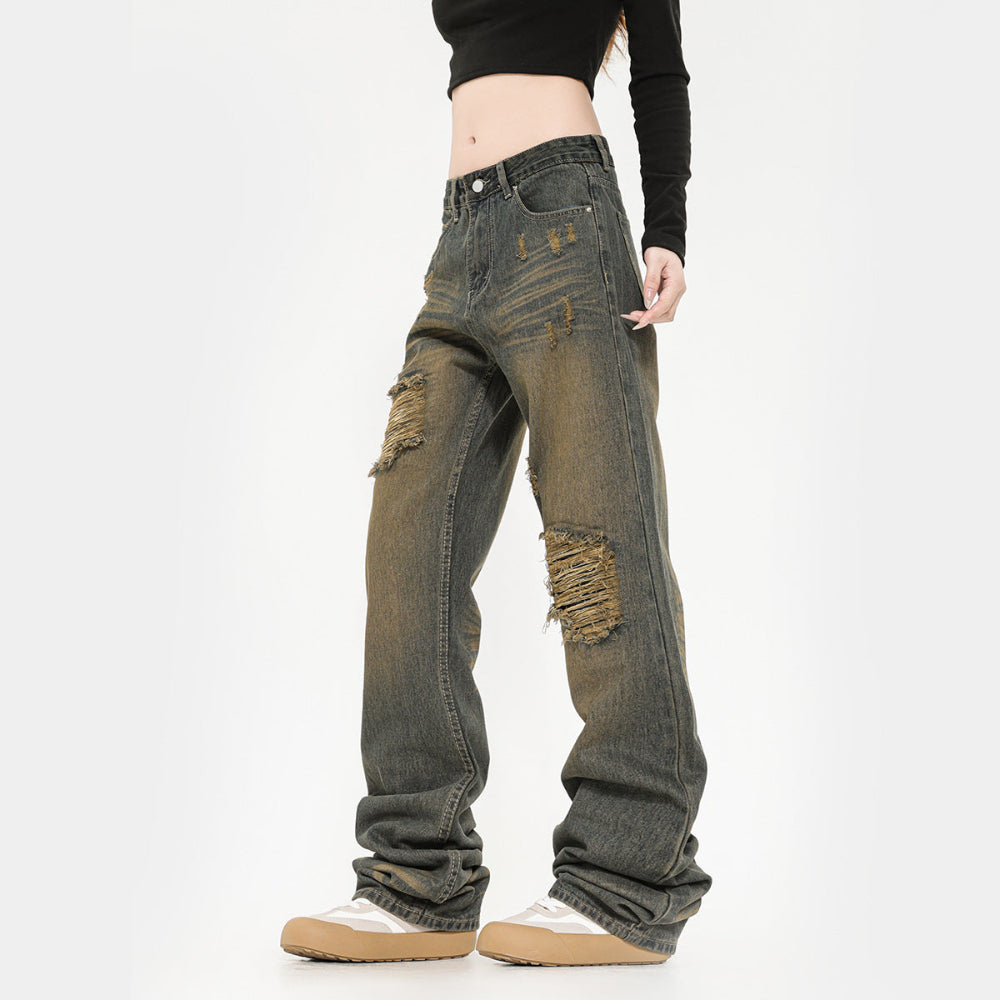 Side view of a model wearing dark greyish-green wide-leg baggy jeans with heavy distressing and large ripped holes with frayed edges on the thighs and knees. The jeans feature a relaxed, oversized fit that stacks at the ankles, paired with a black long-sleeve crop top and chunky white sneakers.