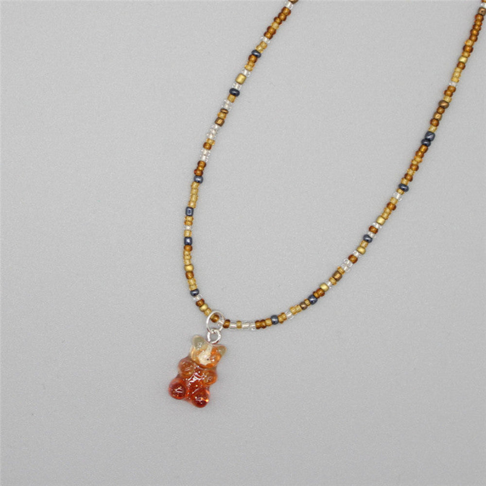 Earthy beaded necklace featuring warm tones of golden brown, amber, navy, and white seed beads with metallic accents. It includes a translucent orange gummy bear pendant. The playful single-strand necklace is laid flat on a light gray background, showcasing its vibrant and whimsical style.