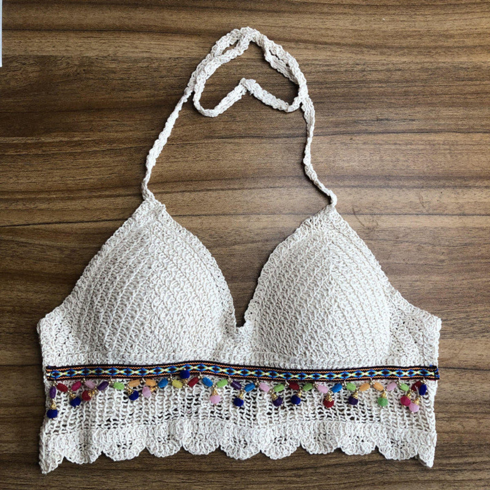 White crochet halter crop top with colorful pom pom and woven trim detail, laid flat on wooden surface. Boutique Jolie boho summer apparel.