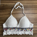 White crochet halter crop top with colorful pom pom and woven trim detail, laid flat on wooden surface. Boutique Jolie boho summer apparel.