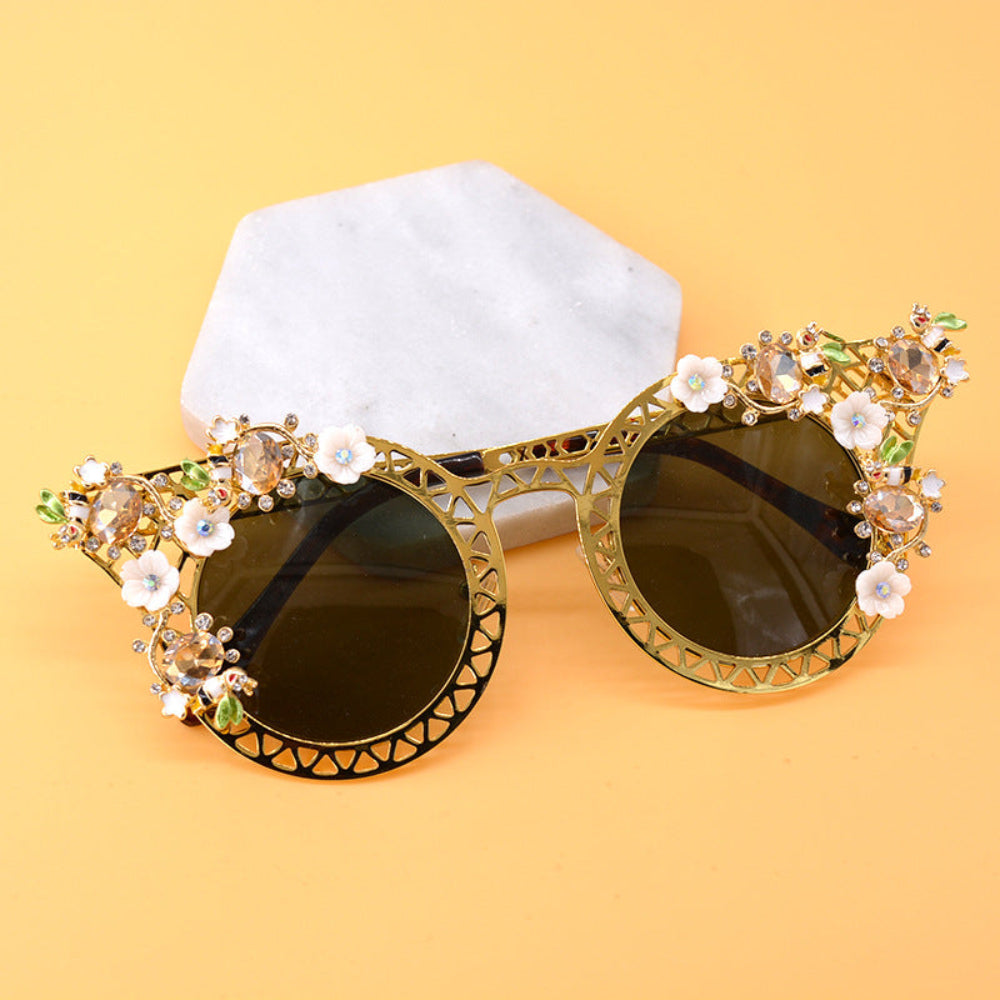 Elegant gold metal cat-eye sunglasses featuring intricate lattice frames, white floral accents, green leaves, sparkling crystals, and dark green-brown tinted lenses, displayed on marble