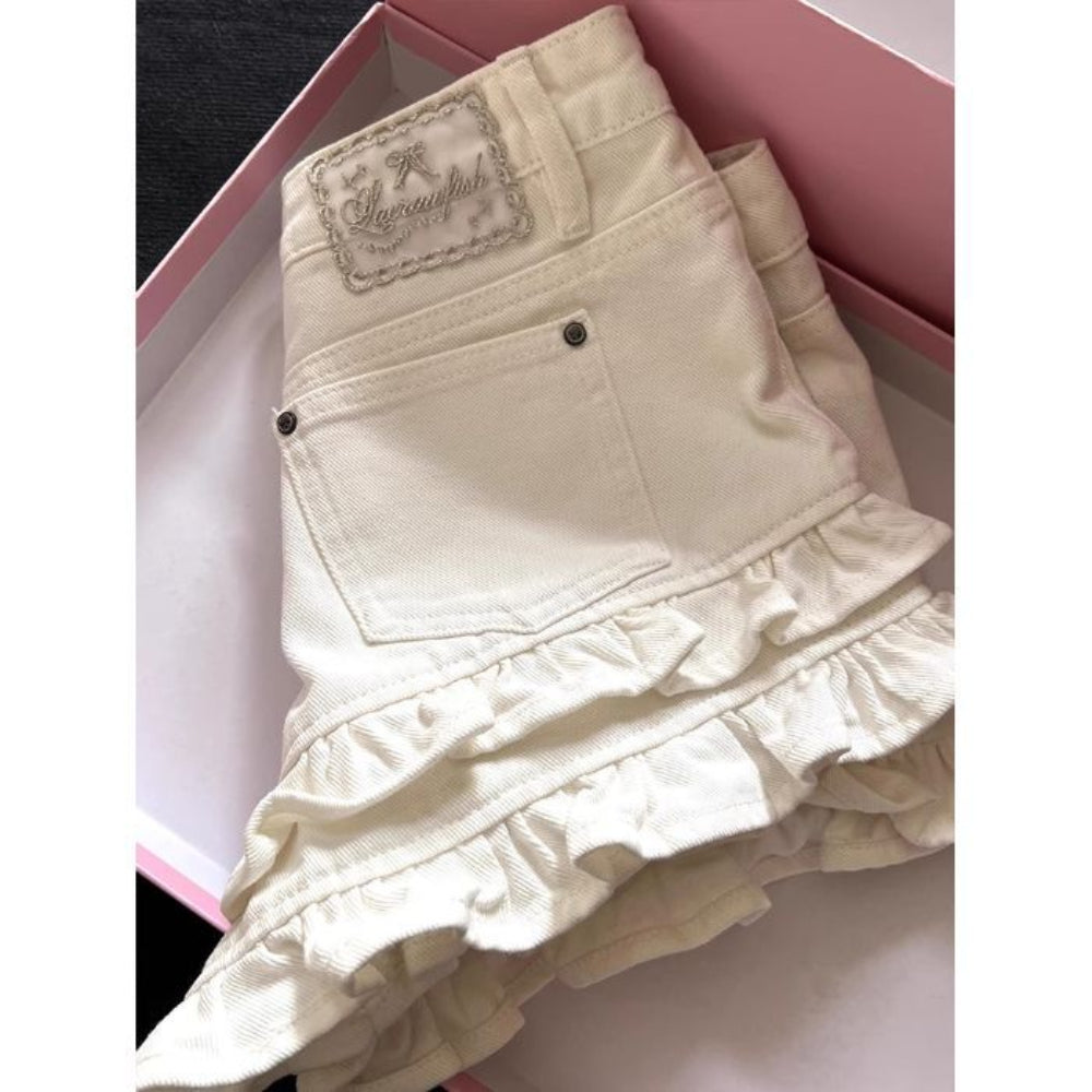 Close-up back view of cream-colored denim shorts with ruffled tiered hems, back pocket, and embroidered 'Sugarwish' brand label, presented inside a pink gift box. Cute feminine fashion detail for Boutique Jolie.