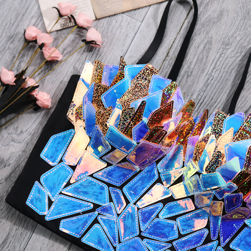 Black tote bag decorated with vibrant iridescent holographic shattered mirror fragments in blue, purple, pink, and gold tones. Features a bold 3D mosaic design with reflective glitter pieces, displayed on a wooden surface with pink flowers.