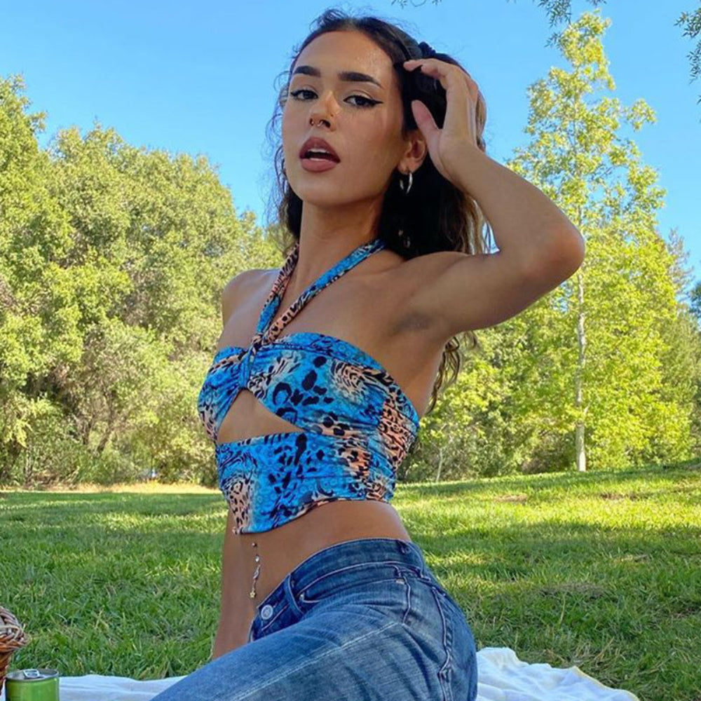 Woman in a blue leopard print halter crop top with twist-front cut-out detail and chain accessory, paired with blue denim jeans. Posing outdoors in a sunny park with green grass and trees, hand touching hair.