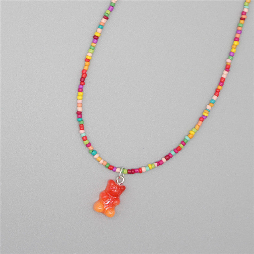 Rainbow beaded necklace featuring a vibrant mix of multicolored seed beads and a single translucent orange gummy bear pendant. Playful single-strand design laid flat on a light gray background, highlighting its fun and cheerful style.