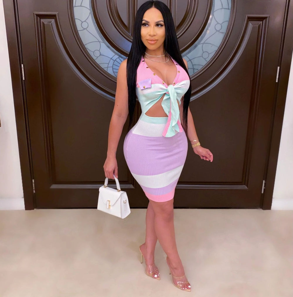 Model in a form-fitting pastel striped dress featuring horizontal blocks of pink, light blue/mint, and purple with a sweetheart neckline and matching bow accent at the waist. Full-body fashion pose holding a white handbag in a stylish indoor setting with an ornate dark door in the background.