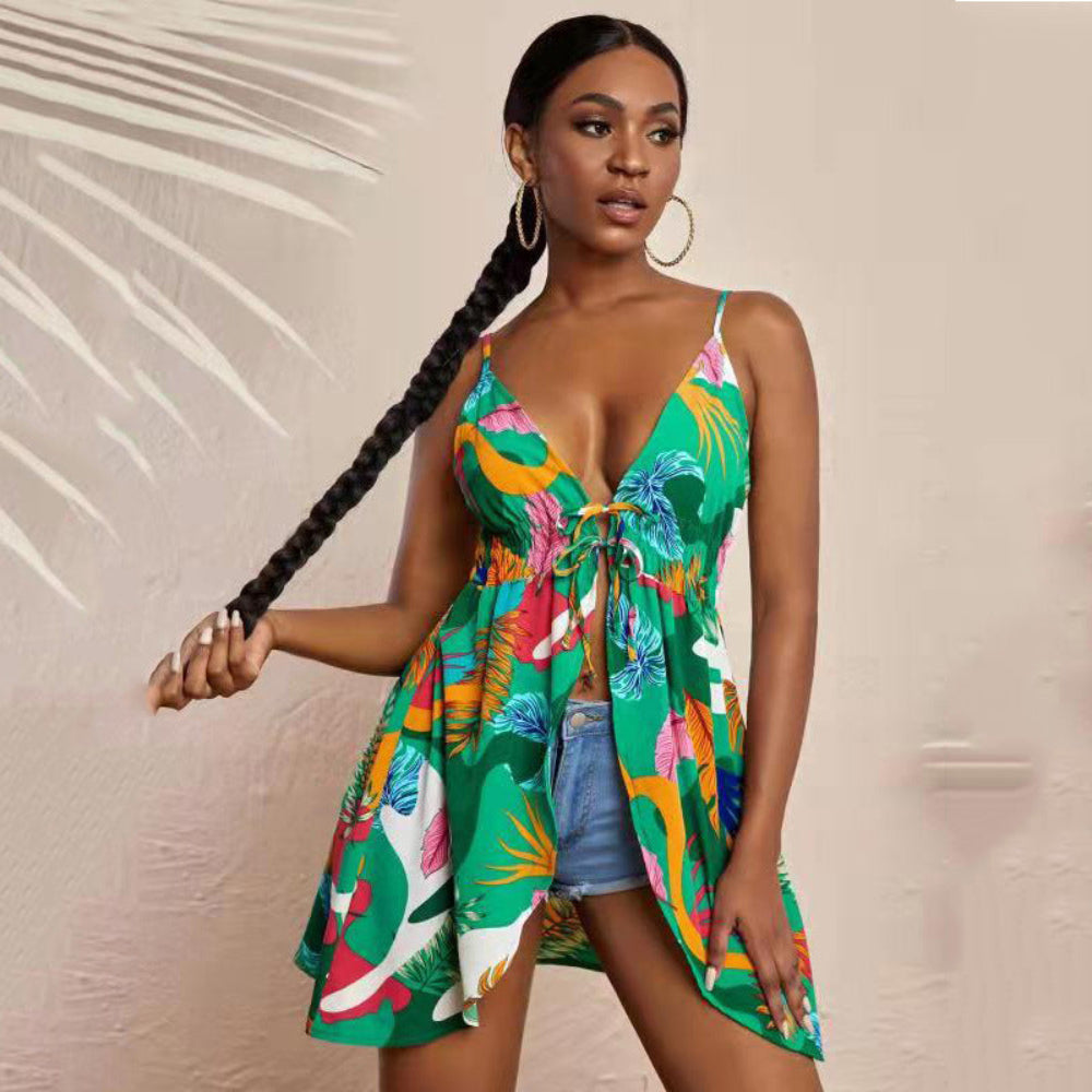 Model wearing a flirty sleeveless tropical print mini dress featuring bold palm leaves and colorful floral patterns on a bright green base. Deep V-neck with front tie detail and flowy hem, styled with denim shorts, large hoop earrings, and long braid, in a stylish studio setting with palm leaf shadow on the wall.