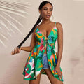 Model wearing a flirty sleeveless tropical print mini dress featuring bold palm leaves and colorful floral patterns on a bright green base. Deep V-neck with front tie detail and flowy hem, styled with denim shorts, large hoop earrings, and long braid, in a stylish studio setting with palm leaf shadow on the wall.