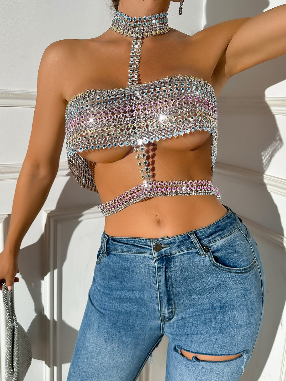 Woman wearing a dazzling iridescent rhinestone halter crop top with high choker neck, multicolored crystal embellishments, and sparkling sequin details. Styled with light blue ripped jeans, showcasing the intricate design under bright lighting.