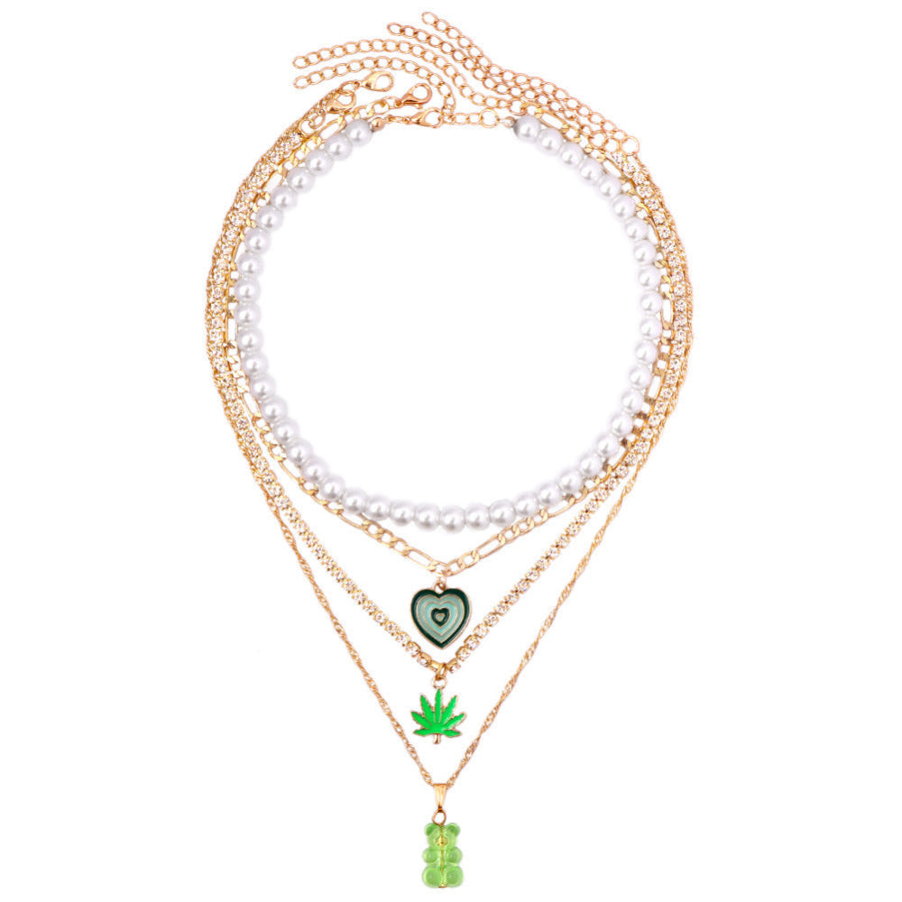 Flat lay of a jewelry stack on soft white fabric featuring a long white pearl necklace, multiple gold chains (including a tennis chain), a green heart-shaped pendant, a green cannabis leaf charm, and a translucent green gummy bear pendant. Elegant product styling highlighting the mixed-metal and playful charm details.