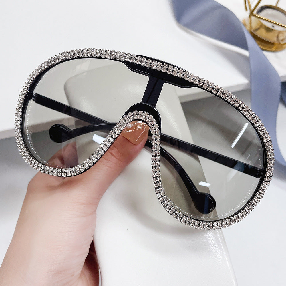 Hand holding oversized clear-gray shield sunglasses with crystal rhinestone trim.