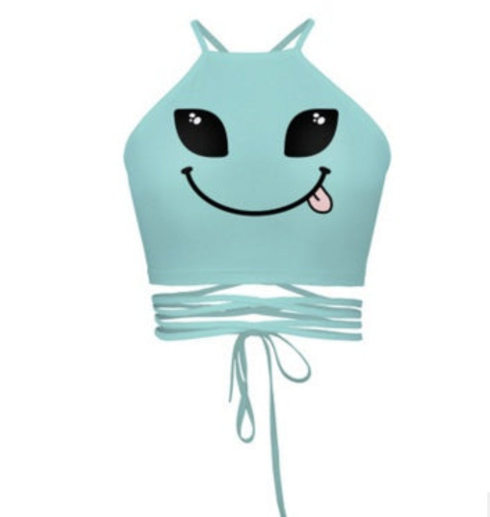 Light blue alien smiley face halter crop top with tongue-out graphic and multi-strap tie-back detail, shown flat lay on white background.