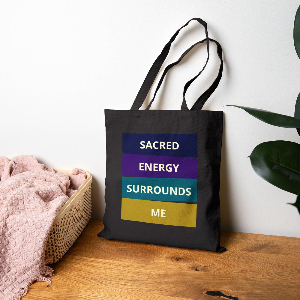 Black canvas tote bag with "SACRED ENERGY SURROUNDS ME" spiritual quote in colorful stacked blocks (navy, purple, teal, gold), styled on wooden surface with pink blanket and plant.
