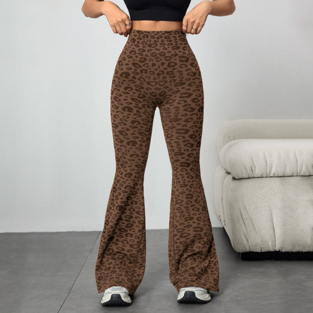 Woman modeling brown leopard print high-waisted flared pants, standing and adjusting the waistband. Paired with a black crop top and sneakers in a bright indoor setting with a gray couch.