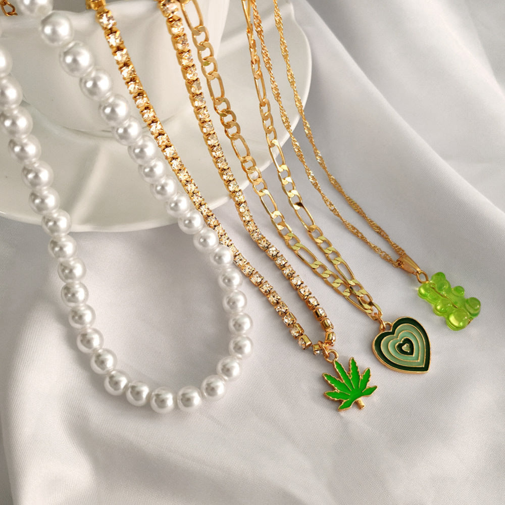 Flat lay of a jewelry stack on soft white fabric featuring a long white pearl necklace, multiple gold chains (including a tennis chain), a green heart-shaped pendant, a green cannabis leaf charm, and a translucent green gummy bear pendant. Elegant product styling highlighting the mixed-metal and playful charm details.