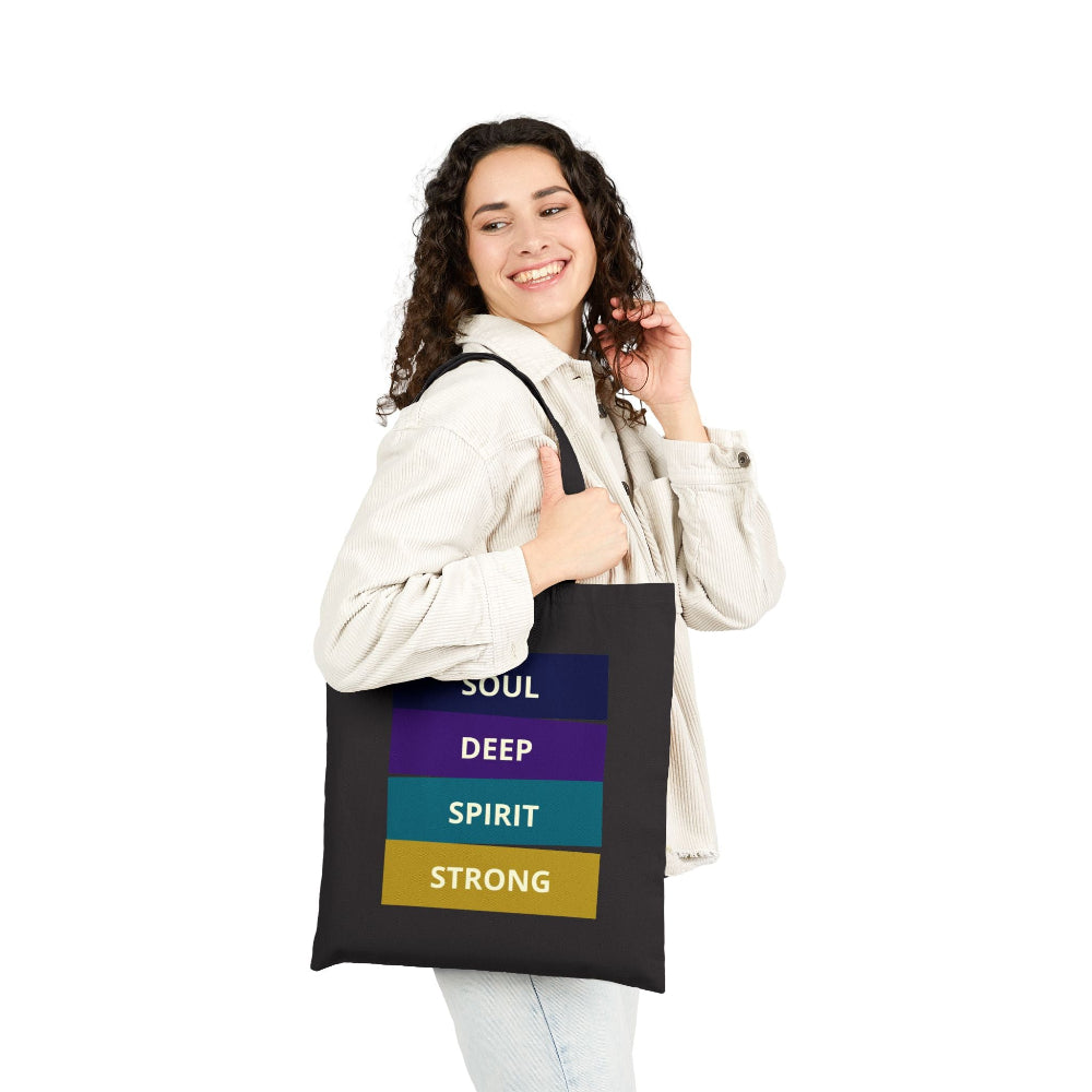 Smiling woman with curly hair wearing a black canvas tote bag with "SOUL DEEP SPIRIT STRONG" motivational quote in colorful stacked blocks, shown against white background.