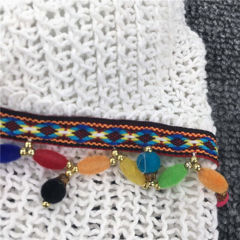 Close-up detail of colorful woven trim with multicolored pom poms and gold beads on white crochet fabric.
