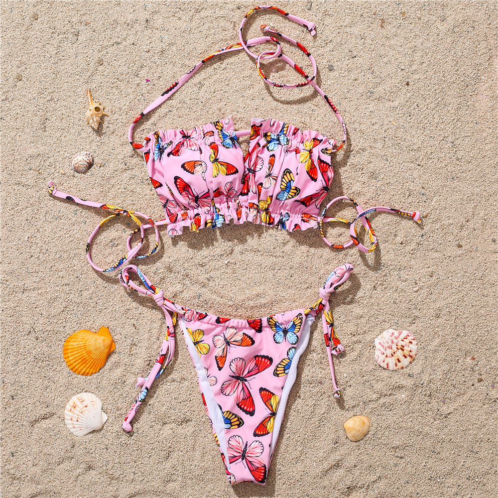 Pink butterfly print bikini on sand – trendy colorful ruffled swimwear set with ties, beach product flat lay.