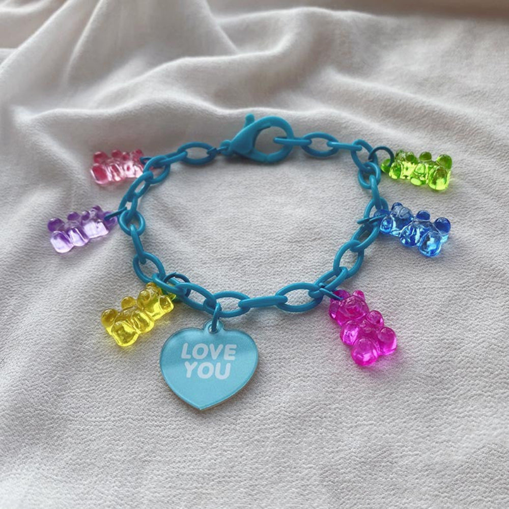Turquoise gummy bear charm bracelet with a bright turquoise chain, featuring colorful translucent gummy bear charms in rainbow shades and a light blue heart-shaped pendant engraved with "LOVE YOU", displayed on white fabric.
