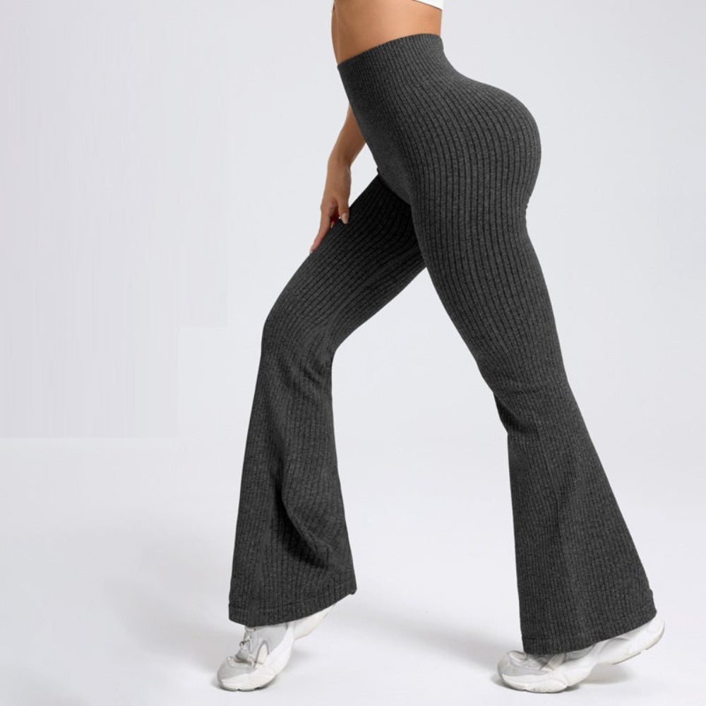 Woman modeling dark gray ribbed knit high-waisted flared pants, shown from a rear three-quarter view. The pants feature a form-fitting silhouette through the hips and thighs that flares out at the hem, paired with white sneakers against a clean white studio background.
