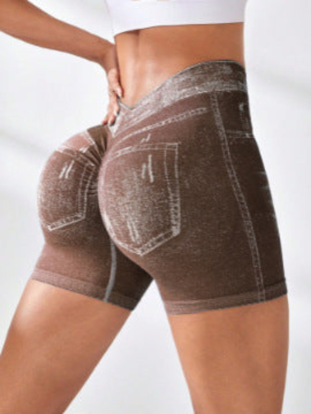 Woman modeling brown denim high-waisted shorts with white contrast stitching and back pockets, shown from a rear three-quarter view against a clean white studio background.